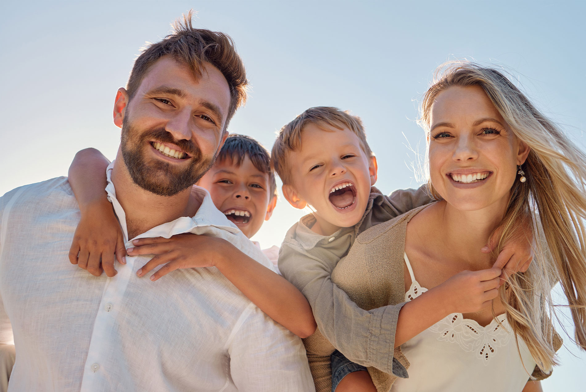 In the image, a man and woman are standing close together, smiling at the camera. They are holding two children who appear to be laughing or enjoying themselves. The family is outdoors during daylight with clear skies in the background.