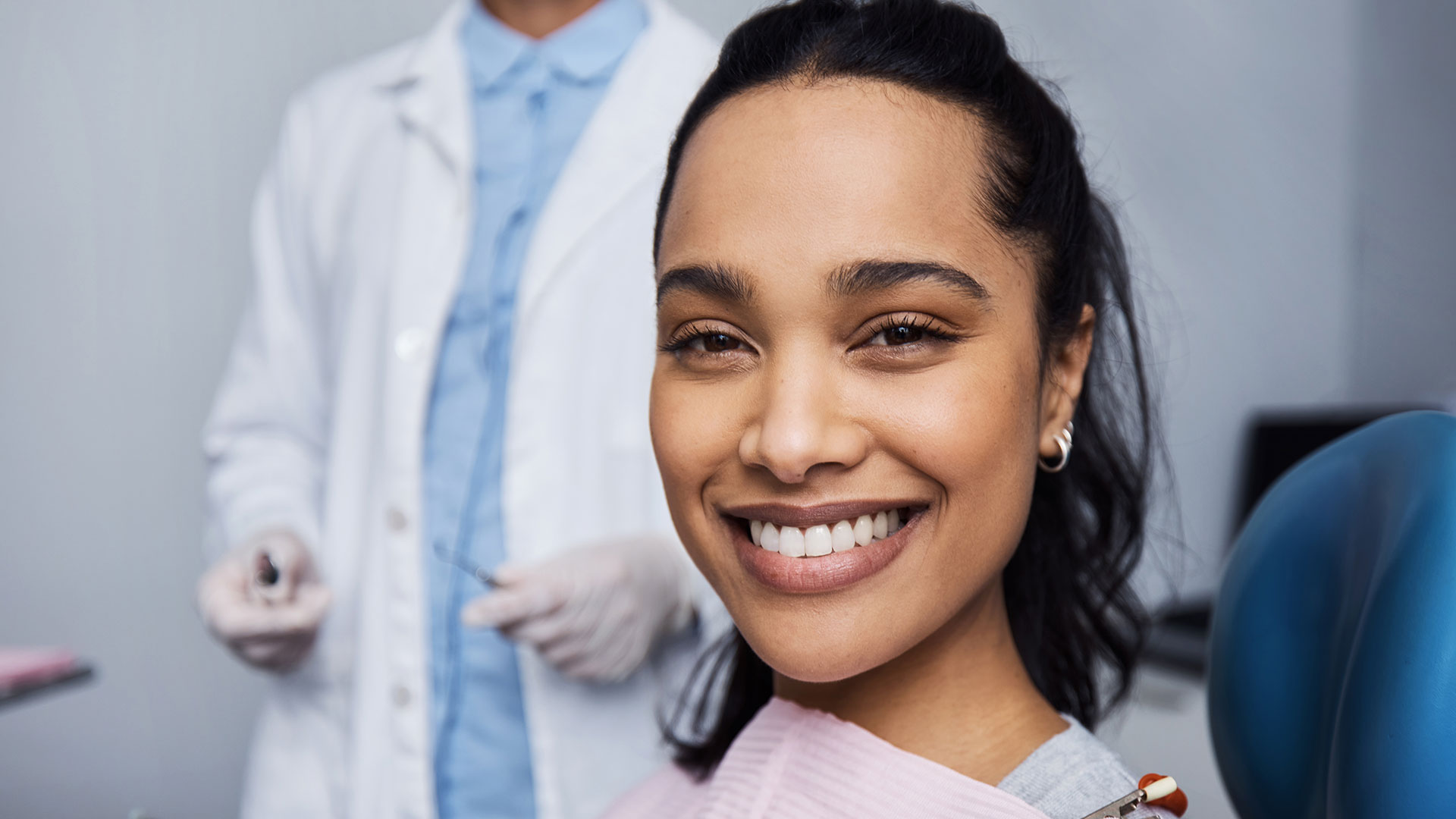 A smiling woman in a dental office, seated in the chair, with a dentist standing behind her.