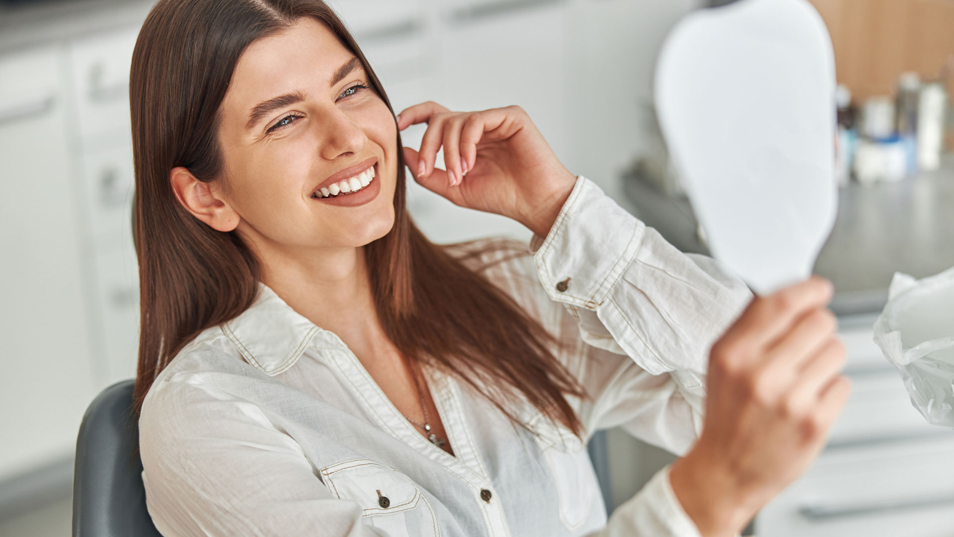 The image shows a woman sitting in front of a mirror, smiling and looking at her own reflection with one hand on her face. She is indoors with a clean, modern aesthetic around her.