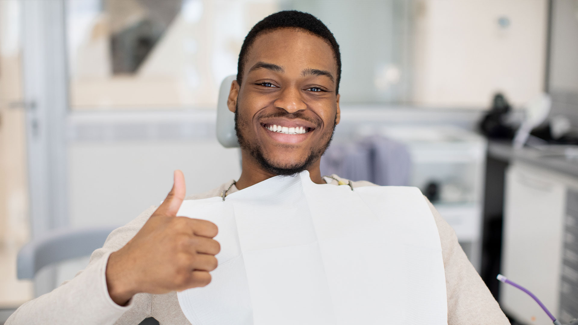 The image shows a person, likely male, sitting in front of a mirror and giving a thumbs-up gesture. He is wearing a surgical mask and appears to be in a dental or medical setting.