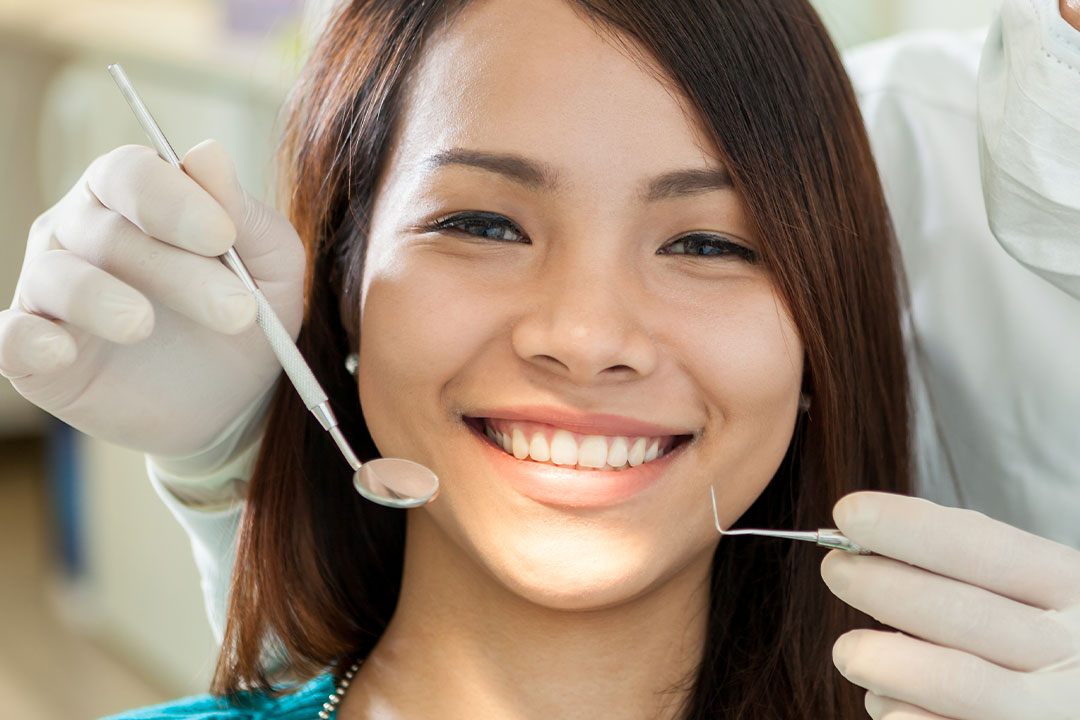 A woman is smiling at the camera, seated in a dental chair with her mouth open, receiving dental care with a dentist wearing gloves standing behind her.