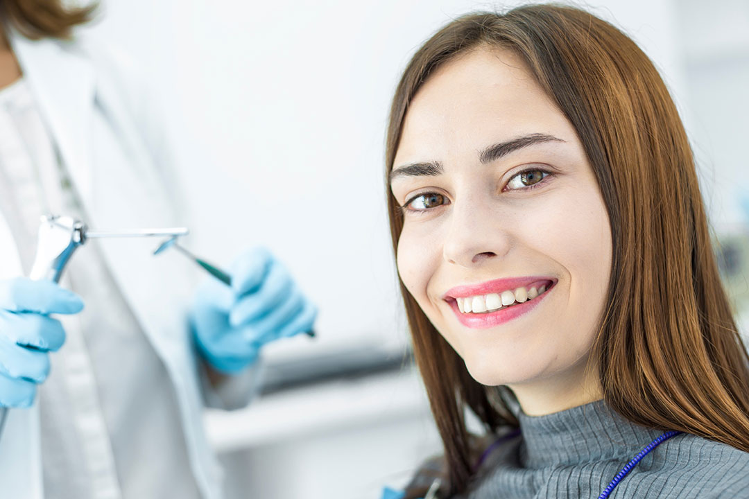 A woman in a dental office, smiling at the camera while seated in a dental chair, with a dentist in protective gear behind her.