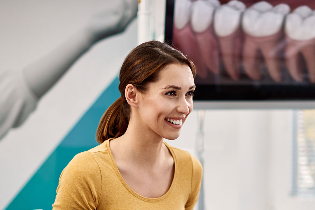 A smiling woman in a yellow top stands in front of a dental office sign, with a wall displaying dental images behind her.