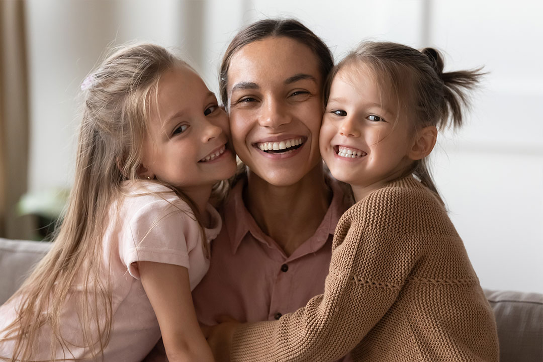An adult woman is smiling and hugging two young children, all of them appear to be indoors.
