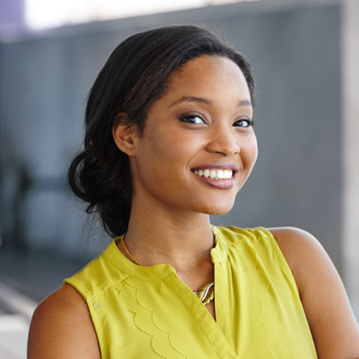 A smiling woman with dark hair, wearing a yellow top and standing against a blurred background.