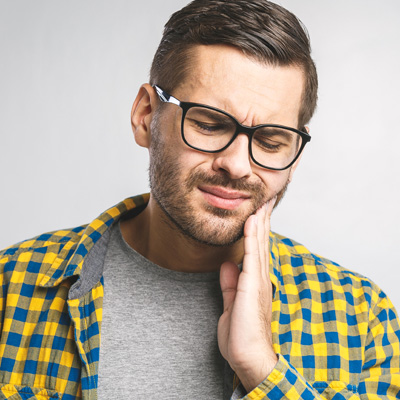 The image shows a man with glasses, wearing a yellow plaid shirt, holding his hand to his mouth in a thoughtful or pensive gesture.