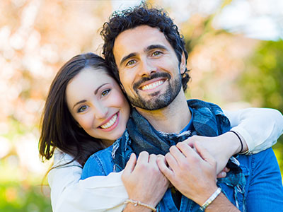 The image shows a man and woman hugging each other, both smiling, with the man wearing a bandana around his neck and the woman wearing a white top. They appear to be in an outdoor setting during the daytime.