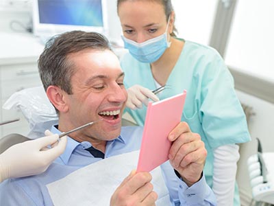 An image featuring a man in a dental chair, holding up a pink card with a surprised expression, while a dentist and dental assistant work on his teeth.