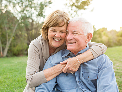 A couple, an older man and woman, embrace warmly in a park setting.