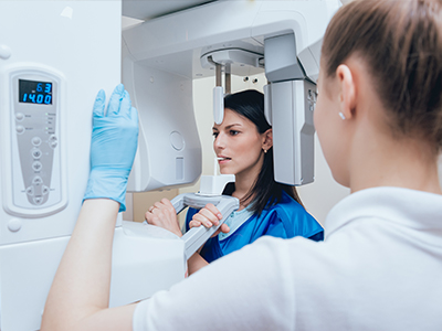 A medical professional in a blue coat using a CT scan machine, with a woman wearing gloves and a face mask observing the display screen.