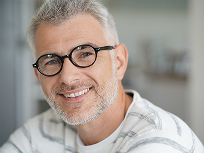 The image is a photograph of a smiling man with gray hair, wearing glasses and a white shirt, looking directly at the camera.