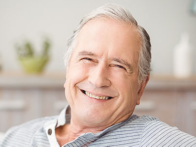 The image shows a man with grey hair, wearing a blue shirt, smiling and looking directly at the camera. He appears to be sitting in a relaxed posture, possibly in a home setting.