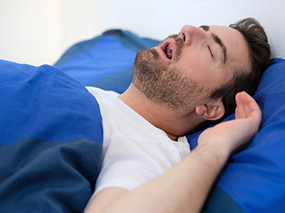 A man is lying in bed, appearing to be asleep with his mouth open.