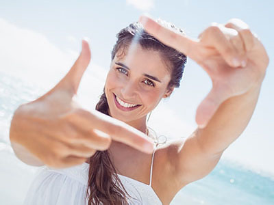 A woman taking a selfie with her hand while smiling at the camera, set against a beach backdrop.